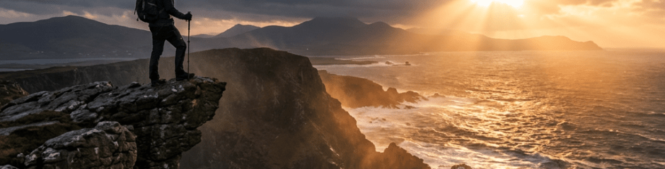 Hiker with backpack standing on rocky cliff overlooking ocean with sunset and clouds